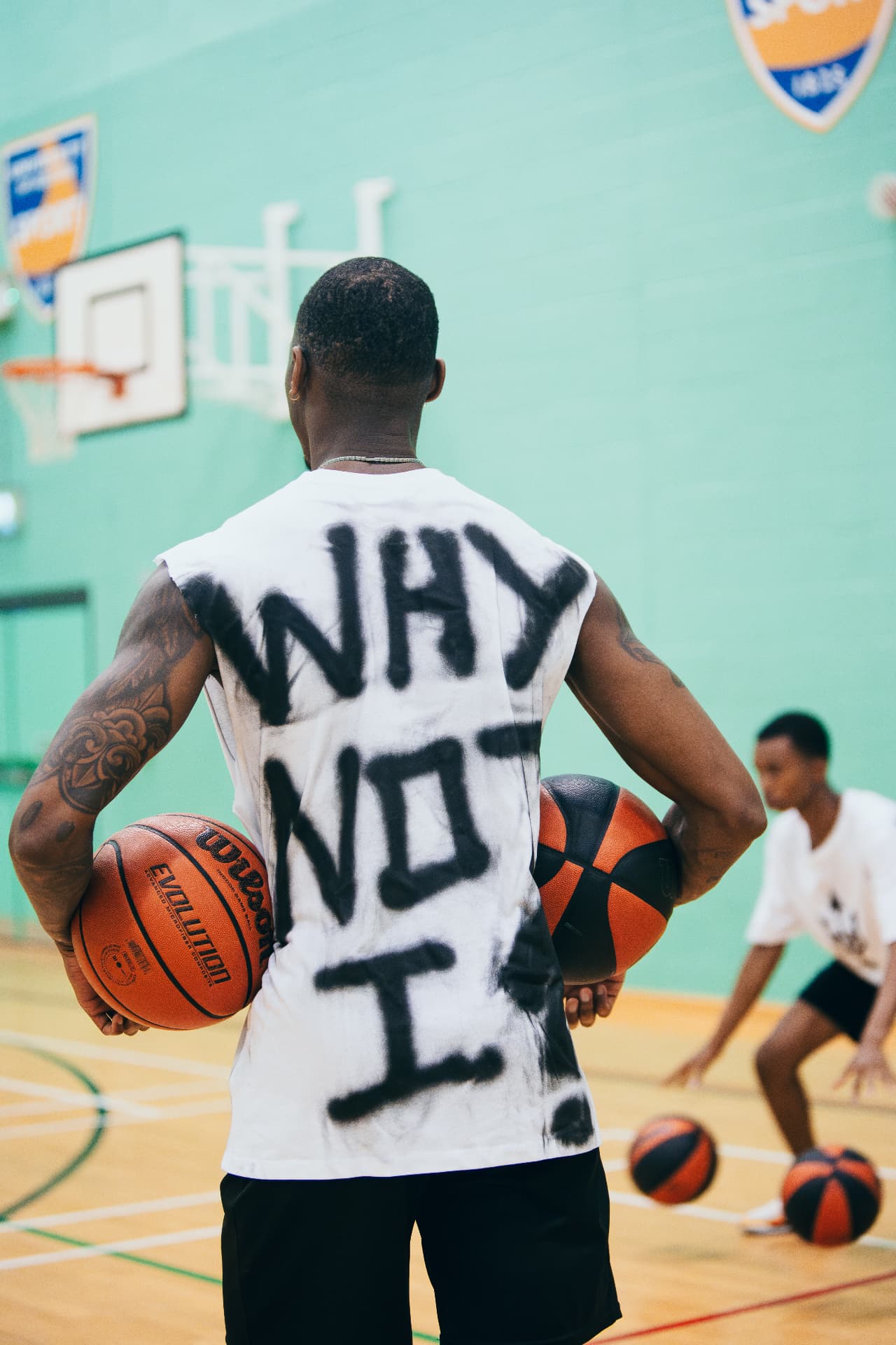 Kofi holds two basketballs behind his back during the first Why Not I Workout in 2024, wearing a Why Not I vest as a young player trains behind him